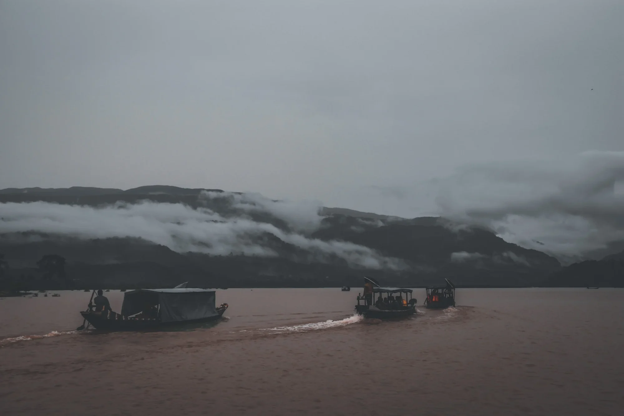 Three covered boats crossing a wide river beneath cloud lying along the hills