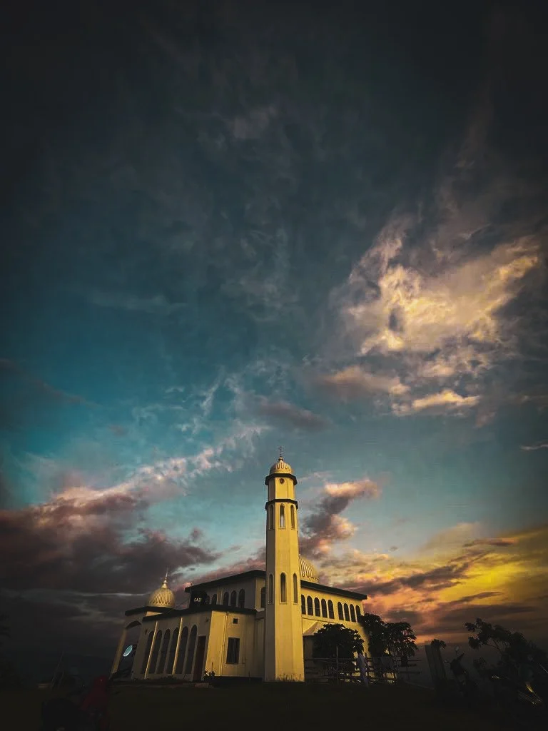 A small mosque and minaret under a wide sky breaking into colour