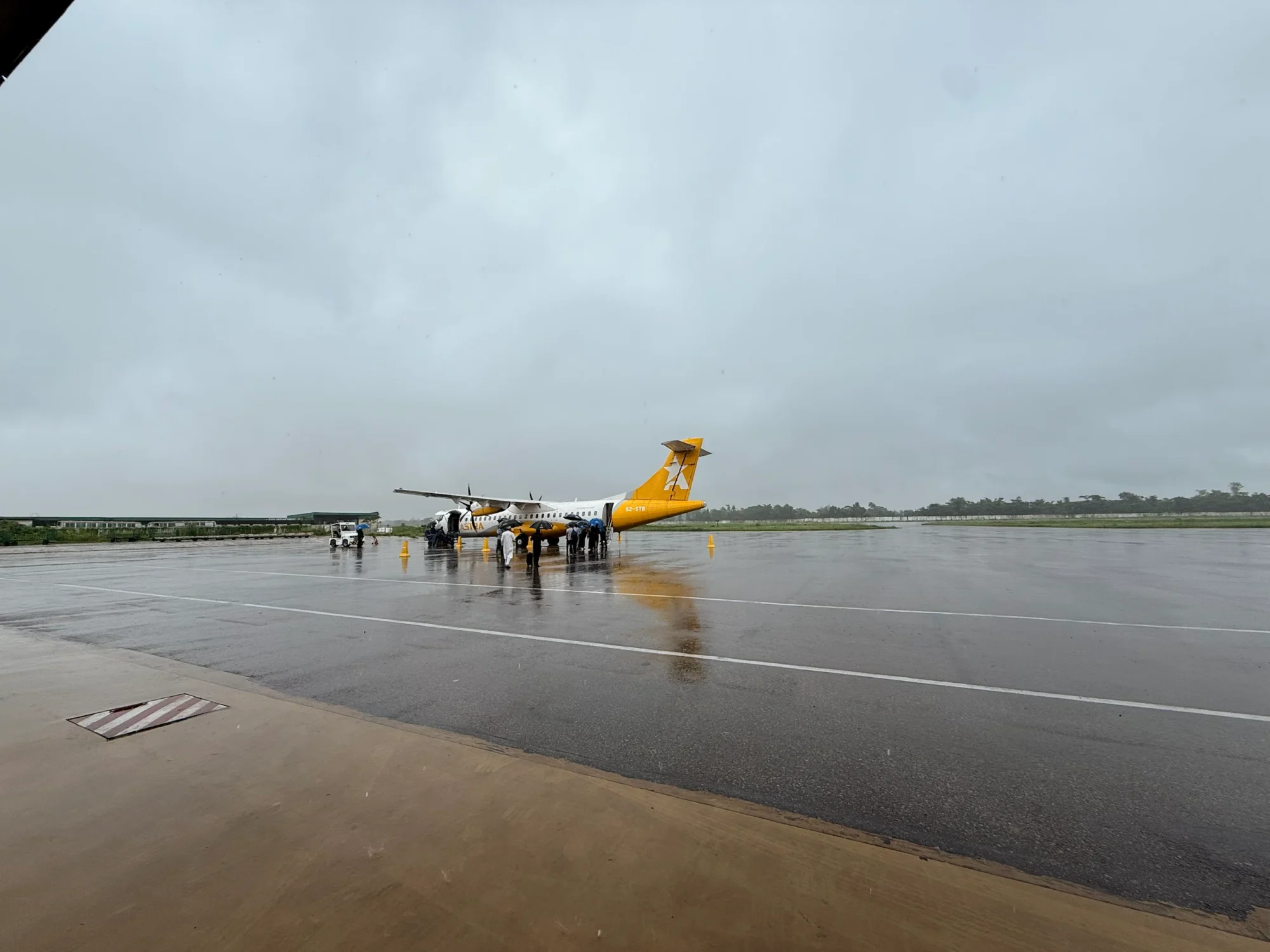 A turboprop aircraft on a wet apron under grey cloud, passengers boarding under umbrellas