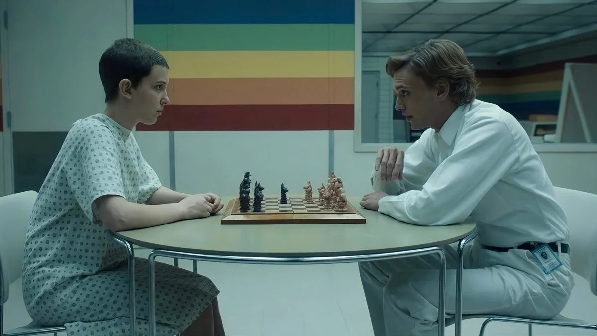 Eleven sitting across a chess board from a man in white in a laboratory
